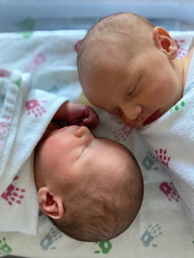 Katie's newborn twins. They're wrapped up to their necks in white hospital blankets, decorated with tiny handprints in various colors. The infants are facing each other with their eyes closed. We can see one twin's tiny hand poking out from underneath the blanket.