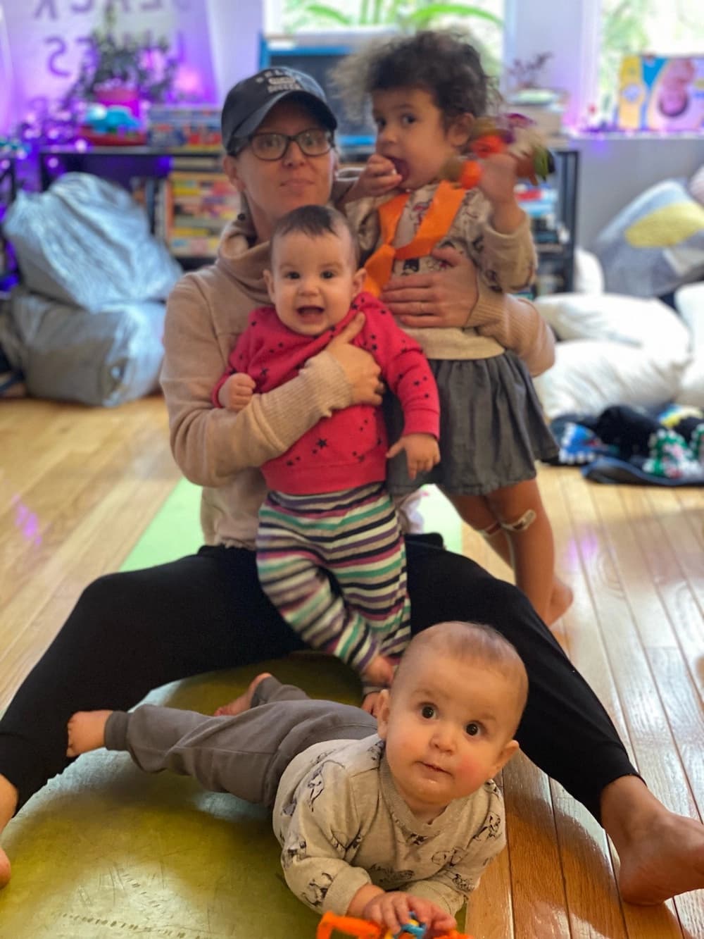 Katie, sitting on the floor at home, her legs out in front of her. Her left arm wraps around her toddler, who looks like she's eating something. The toddler isn't fully stationary; her left arm is blurry. Katie's right arm is wrapping around one of the twins, helping her to stay 'standing' up. She looks happy. The other twin is in front of Katie, on his tummy, looking up at the camera.