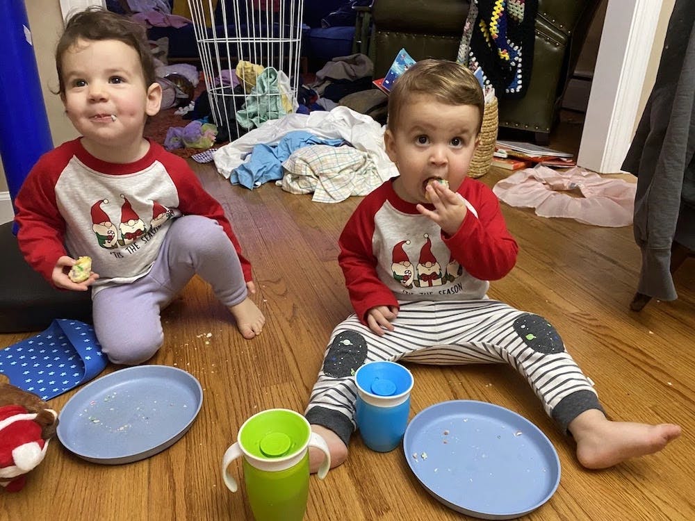 Katie's twins, around two years old. They're sitting on the dining room floor, at home, eating dinner. They're wearing matching shirts: some elves in red hats with the text 'tis the season' underneath. Their empty plates have plenty of crumbs; their sippy cups are in front of them. Twin A (on the left) has food in her hand, and is smiling at someone who is to the left of the camera. Twin B is actively eating - mid-bite - and he is looking at the camera. The background is messy. We can see a mostly empty hamper and clothes strewn about the ground.