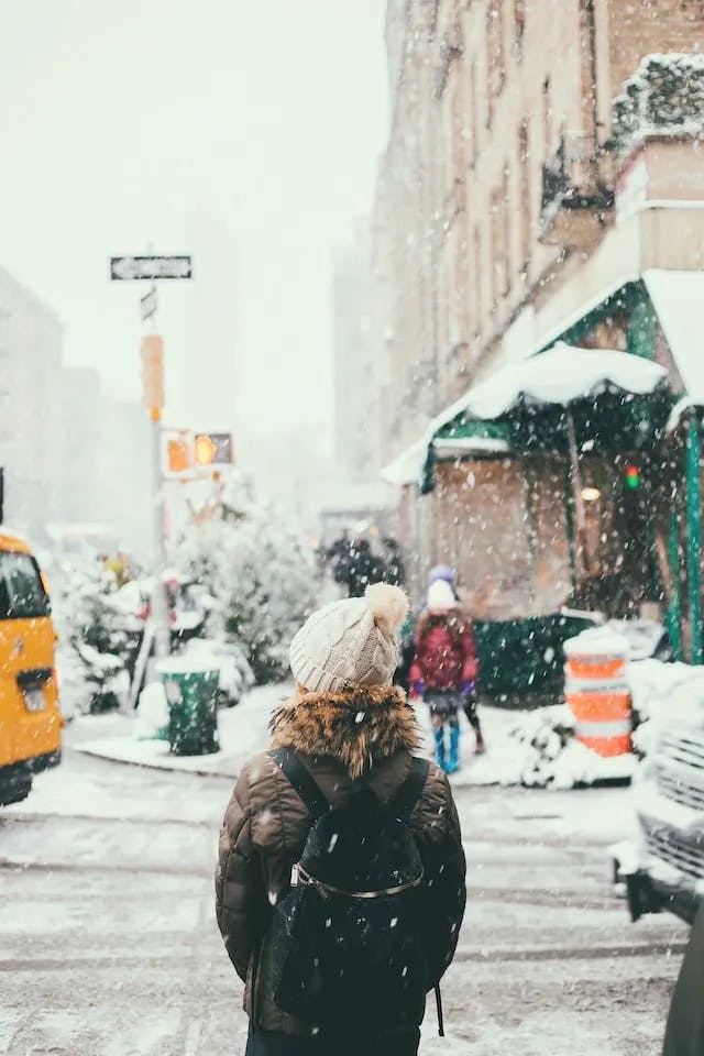Woman wearing a backpack, an off-white hat, and winter jacket with her back facing the camera. She is downtown in an unknown city, waiting to cross a small intersection. There is snow falling and snow on the ground.