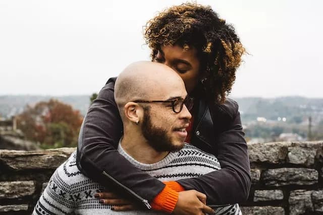 A couple in love. The woman is behind the man, with her arms coming over his shoulders and resting on his chest. Neither of them are looking at the camera. She's kissing the side of his head. He is smiling and his head is turned to the left.