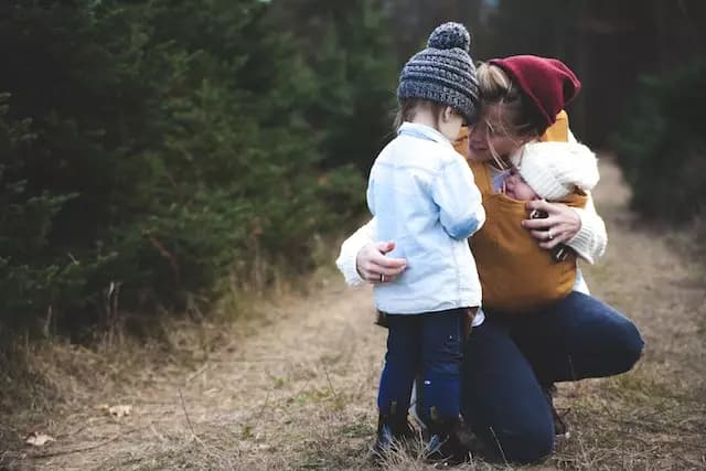 A woman with a baby on her chest, kneeling down and comforting a small child. All are wearing hats. They're on a light brown path and there are evergreen trees in the background.