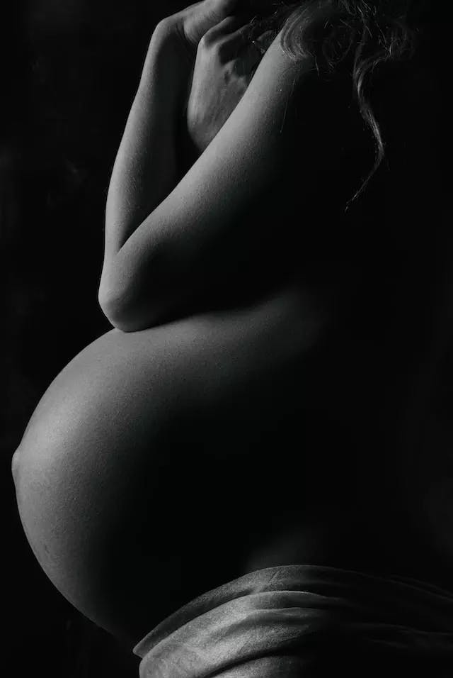 In grayscale, the side view of a heavily pregnant woman. Her belly is the focal point and she wraps her arms around herself, covering her breasts.