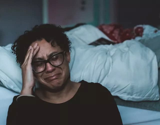 Woman sitting with her back against a bed. She is crying, obviously in distress, and has her hand on her forehead. Her eyes are closed.